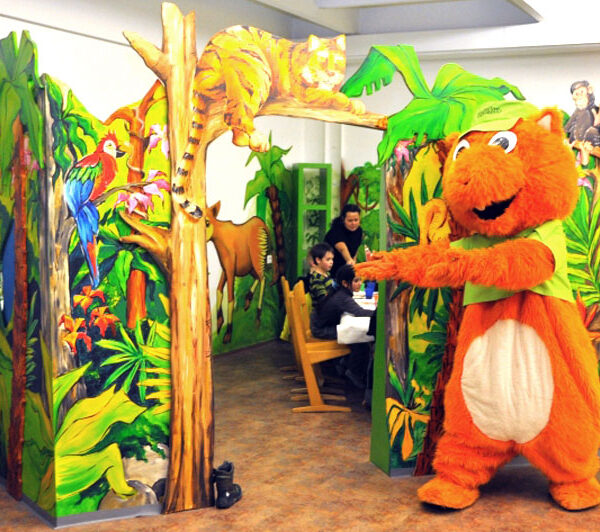 Eine Person in einem orangefarbenen Tierkostüm steht vor einem farbenfrohen, dschungelartigen Spielbereich, in dem Kinder an einem Tisch sitzen.