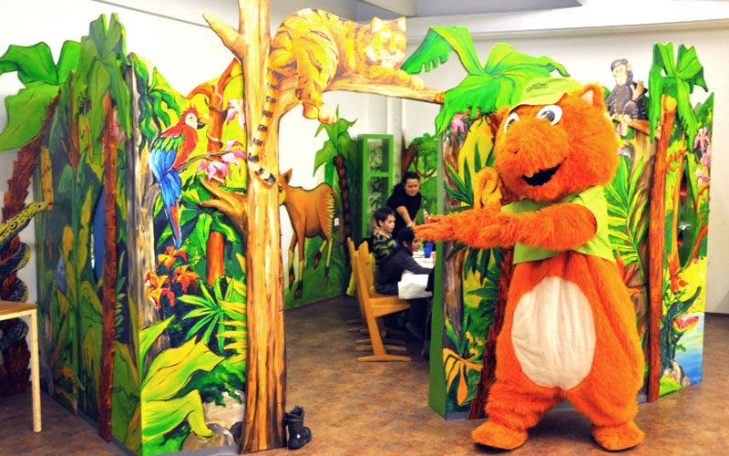 Eine Person in einem orangefarbenen Tierkostüm steht vor einem farbenfrohen, dschungelartigen Spielbereich, in dem Kinder an einem Tisch sitzen.