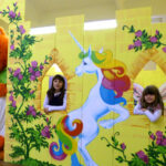 Zwei junge Mädchen posieren hinter einer gelben, mit einem Einhorn geschmückten Schlosskulisse, während eine Person in einem großen orangefarbenen Katzenkostüm in der Nähe steht.