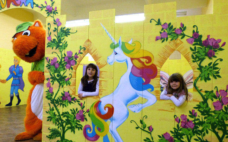 Zwei junge Mädchen posieren hinter einer gelben, mit einem Einhorn geschmückten Schlosskulisse, während eine Person in einem großen orangefarbenen Katzenkostüm in der Nähe steht.