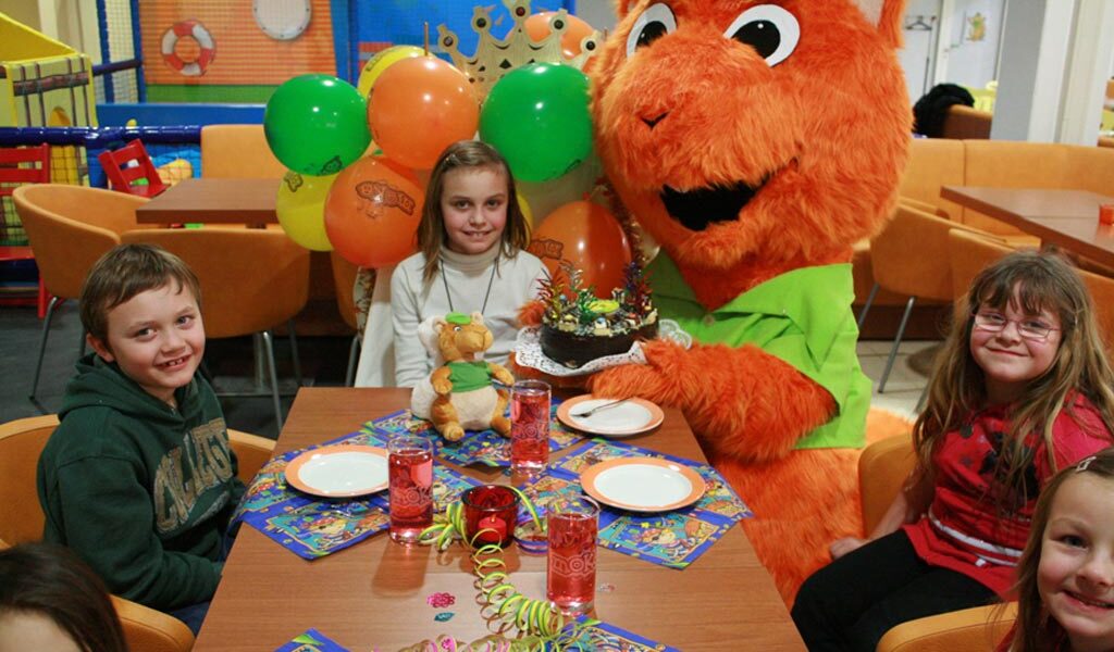 Kinder sitzen um einen Tisch mit einer Torte und Partydekoration, dazu eine Person in einem orangefarbenen Tierkostüm mit Luftballons in einer bunt geschmückten Halle.