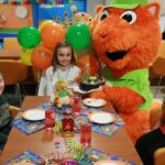 Kinder sitzen um einen Tisch mit einer Torte und Partydekoration, dazu eine Person in einem orangefarbenen Tierkostüm mit Luftballons in einer bunt geschmückten Halle.