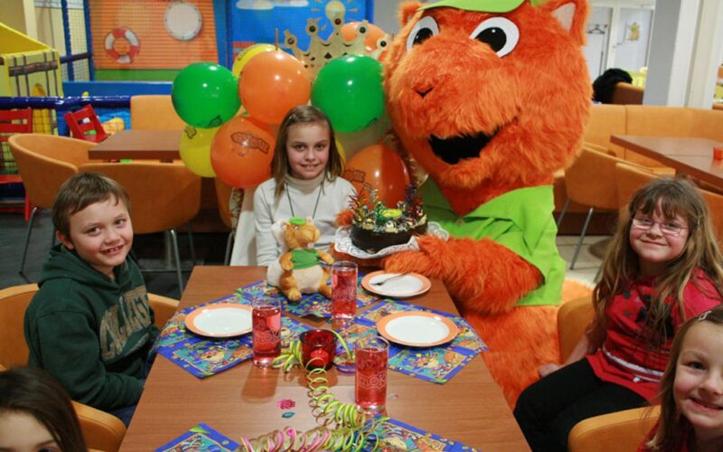 Kinder sitzen um einen Tisch mit einer Torte und Partydekoration, dazu eine Person in einem orangefarbenen Tierkostüm mit Luftballons in einer bunt geschmückten Halle.