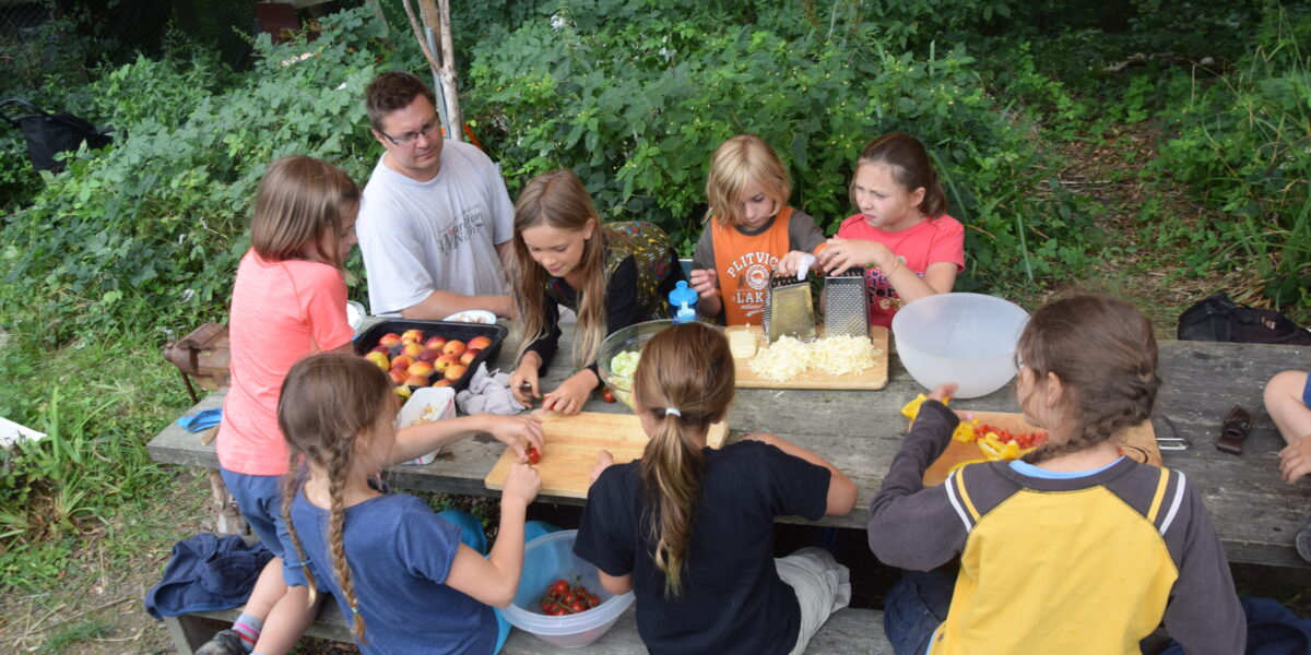 Eine Gruppe von Kindern und ein Erwachsener bereiten gemeinsam Obst und Gemüse an einem Holztisch im Freien zu, der von Grünpflanzen umgeben ist.