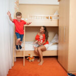 In einem hell erleuchteten, aufgeräumten Schlafzimmer sitzt eine Frau mit einem Baby im Arm auf dem unteren Etagenbett, während ein kleiner Junge von der Leiter des oberen Etagenbetts springt.