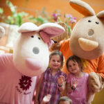 Zwei Kinder stehen zwischen zwei Personen in großen Hasenkostümen, die alle lächeln und in die Kamera winken, in einer hell erleuchteten Innenumgebung.
