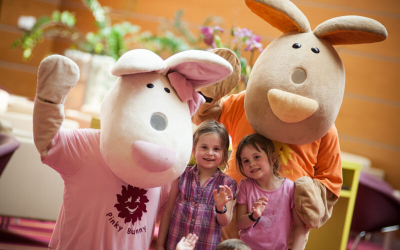 Zwei Kinder stehen zwischen zwei Personen in großen Hasenkostümen, die alle lächeln und in die Kamera winken, in einer hell erleuchteten Innenumgebung.