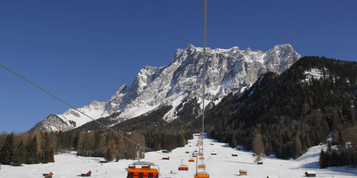 Orangefarbene Skiliftkabinen steigen über ein verschneites Skigebiet mit verstreuten Holzhütten, bewaldeten Hängen und einem felsigen Berg im Hintergrund unter einem klaren blauen Himmel auf.
