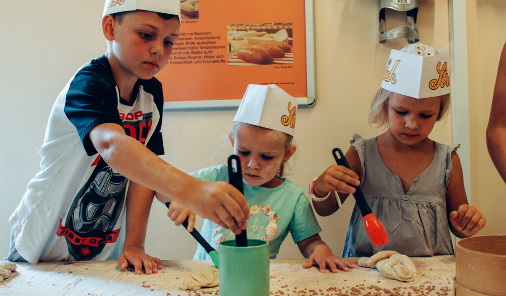 Drei Kinder mit Papierhüten bestreichen an einem Tisch Teig mit Eigelb und nehmen an einer Backaktivität teil.