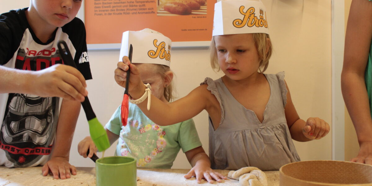 Drei kleine Kinder mit Papierhüten backen gemeinsam und tragen mit Pinseln Flüssigkeit aus einem grünen Becher auf den Teig auf einem mit Samen bedeckten Tisch auf.