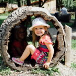 Zwei junge Mädchen sitzen im Freien in einem ausgehöhlten Baumstamm; eines lächelt in die Kamera, während das andere im Schatten sitzt.