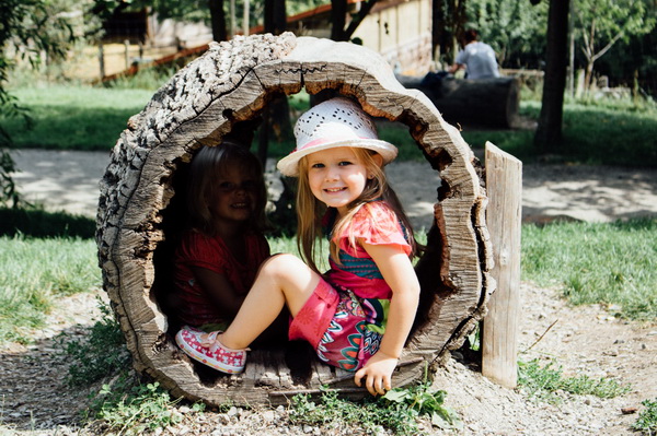 Zwei junge Mädchen sitzen im Freien in einem ausgehöhlten Baumstamm; eines lächelt in die Kamera, während das andere im Schatten sitzt.