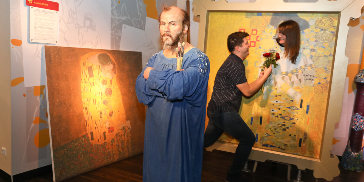 Eine Wachsfigur eines bärtigen Mannes steht vor Gemälden, während im Hintergrund ein Paar als Teil des Kunstwerks posiert.