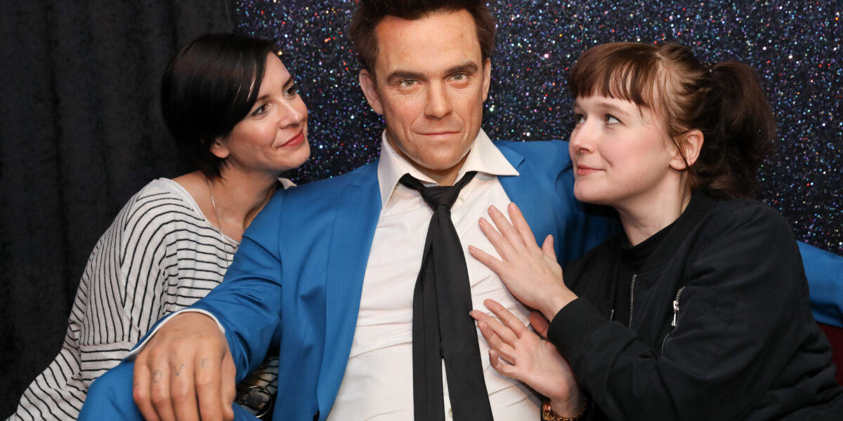 Zwei Frauen posieren mit einer Wachsfigur eines Mannes in einem blauen Anzug vor einem glitzernden Hintergrund.