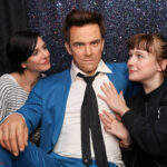 Zwei Frauen posieren mit einer Wachsfigur eines Mannes in einem blauen Anzug vor einem glitzernden Hintergrund.