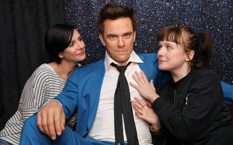 Zwei Frauen posieren mit einer Wachsfigur eines Mannes in einem blauen Anzug vor einem glitzernden Hintergrund.