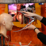 Eine Person misst mit einem Metallmessschieber den Umfang eines Puppenkopfes, wobei Haarfarbmuster und ein orangefarbener Hintergrund zu sehen sind.