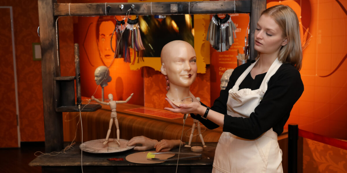 Eine Frau in einer Schürze hält einen Schaufensterpuppenkopf in einer Werkstatt, in der Schaufensterpuppenteile, Werkzeuge und Puppen auf einem Holztisch ausgestellt sind.