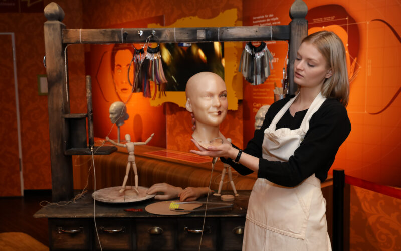 Eine Frau in einer Schürze hält einen Schaufensterpuppenkopf in einer Werkstatt, in der Schaufensterpuppenteile, Werkzeuge und Puppen auf einem Holztisch ausgestellt sind.