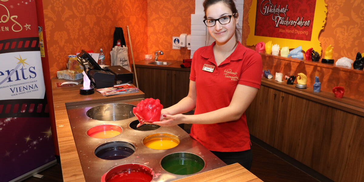 Eine Frau in einem roten Hemd hält eine rote Wachshandform neben einer Station mit Fässern mit farbigem Wachs in einem Wachs-Hand-Tauchstudio.