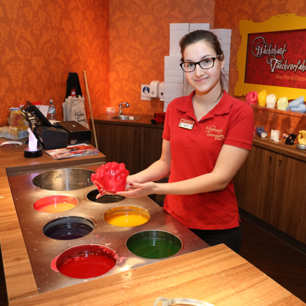 Eine Frau in einem roten Hemd hält eine rote Wachshandform neben einer Station mit Fässern mit farbigem Wachs in einem Wachs-Hand-Tauchstudio.