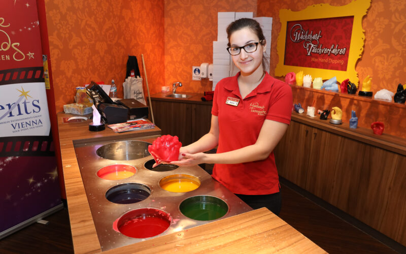 Eine Frau in einem roten Hemd hält eine rote Wachshandform neben einer Station mit Fässern mit farbigem Wachs in einem Wachs-Hand-Tauchstudio.