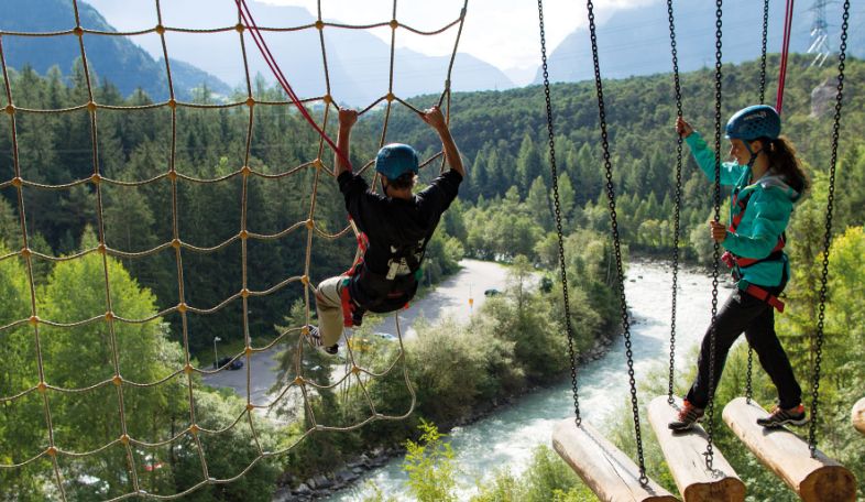 Zwei Personen mit Helmen und Klettergurten navigieren in einem Hochseilgarten über einem Fluss, mit Bäumen und Bergen im Hintergrund.