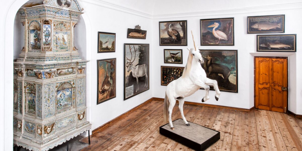 In einem Museumsraum mit Holzfußboden stehen eine lebensgroße Einhornskulptur, Tierbilder an den Wänden und ein kunstvoller Kachelofen in der Ecke.