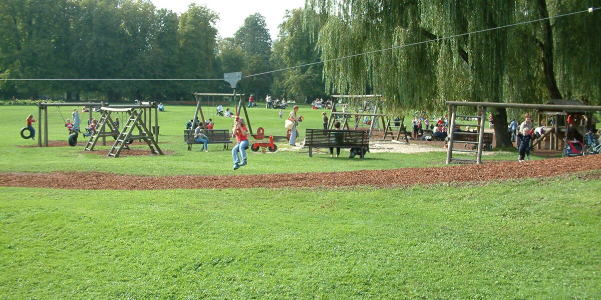Kinder und Erwachsene befinden sich auf einem Spielplatz in einem Park, mit Schaukeln, Rutschen und Klettergerüsten auf einer von Bäumen umgebenen Wiese.
