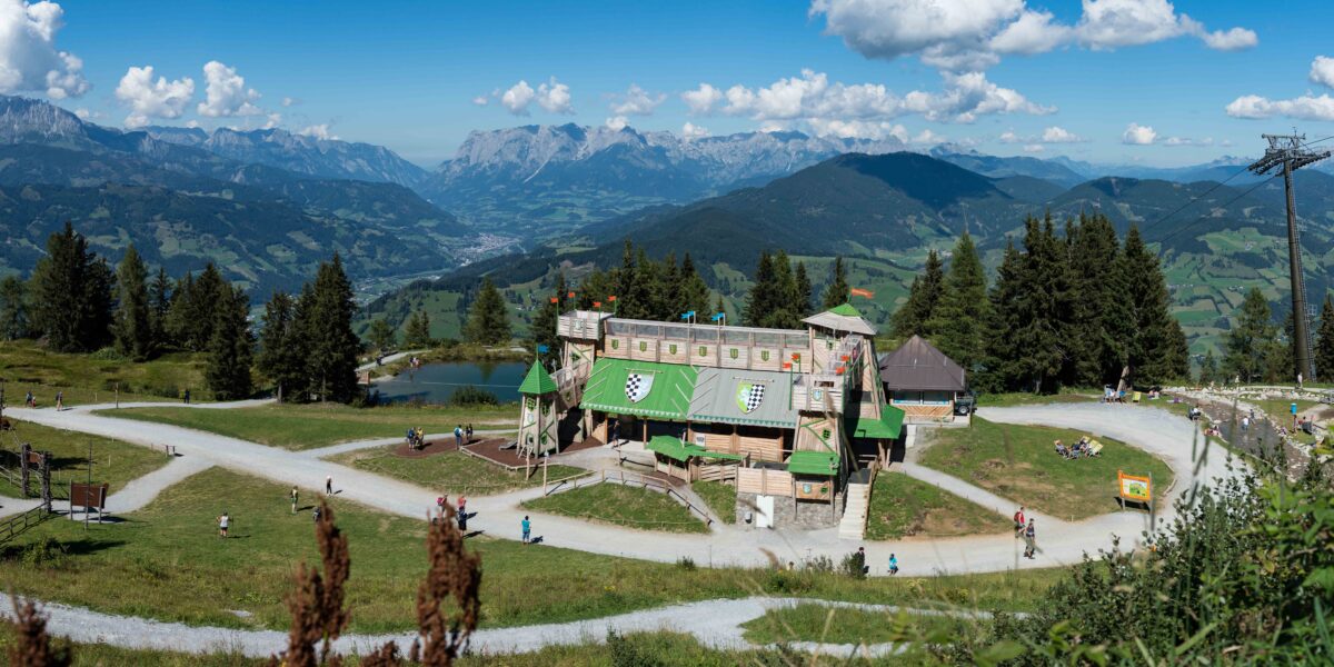 Ein Spielplatz in Form einer Holzburg liegt an einem Berghang, umgeben von Wanderwegen, Bäumen und entfernten Bergketten unter einem blauen Himmel mit vereinzelten Wolken.