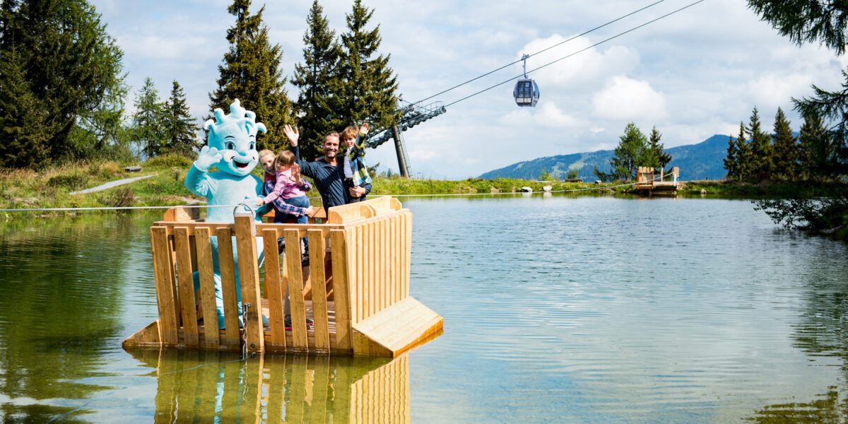 Eine Familie und ein blaues Maskottchen stehen auf einem Holzfloß, das auf einem See schwimmt. Im Hintergrund sind Bäume, Berge und eine Seilbahn zu sehen.