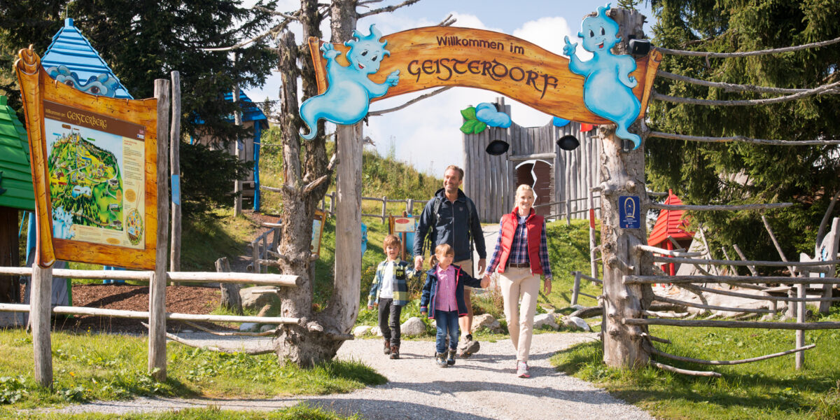 Eine vierköpfige Familie geht durch das Eingangstor von Geisterdorf, einem Themenpark mit verspielten Geisterdekorationen und bunten Gebäuden im Hintergrund.