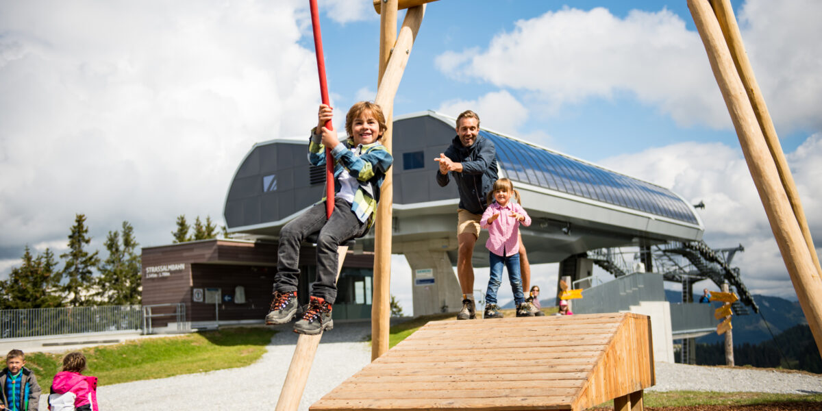 Ein Kind fährt auf einer Seilrutsche, während ein Erwachsener und ein weiteres Kind an einem sonnigen Tag auf einer Holzplattform auf einem Spielplatz in der Nähe einer Skiliftstation stehen.