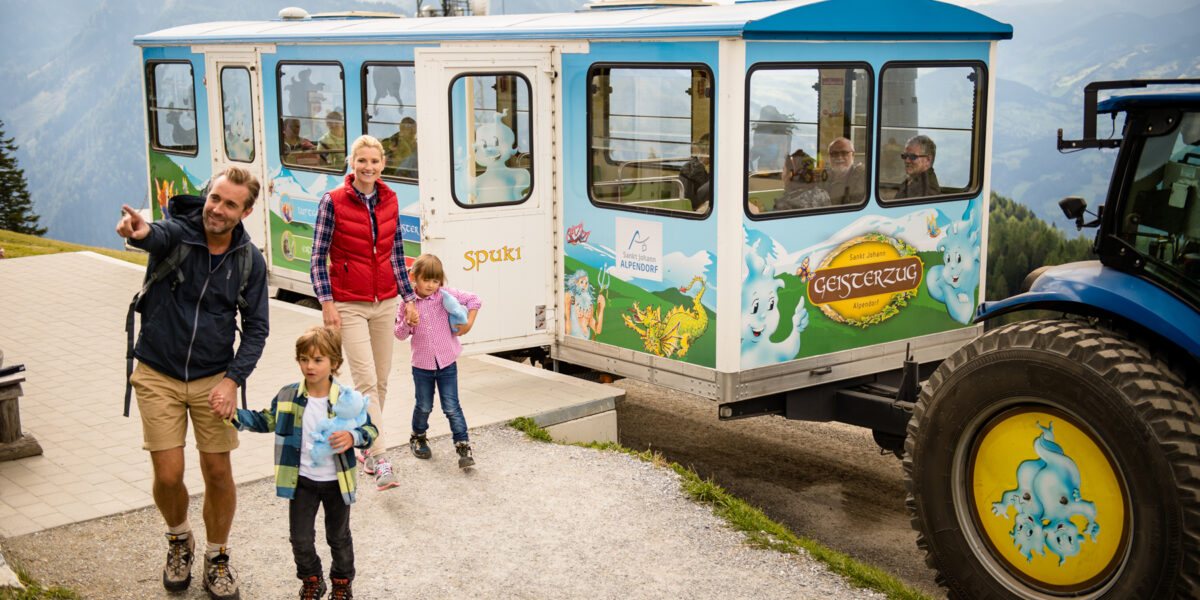 Eine Familie mit zwei Kindern verlässt eine farbenfrohe, mit Cartoon-Geistern geschmückte Bergbahn, mit Bergen und einem blauen Traktor im Hintergrund.
