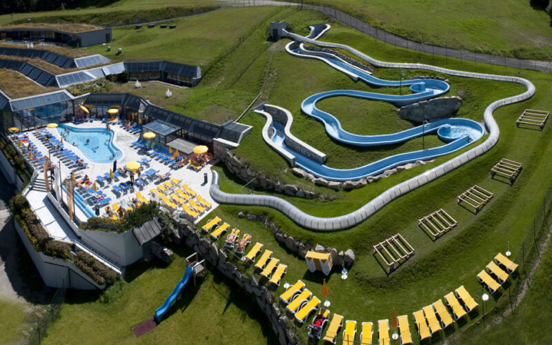 Luftaufnahme eines Wasserparks im Freien mit einem Poolbereich, Sonnenliegen und einer gewundenen blauen Wasserrutsche auf einem grasbewachsenen Hügel.