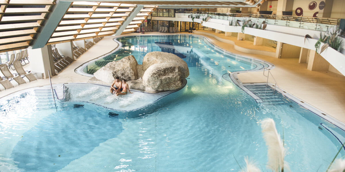 Hallenbad mit klarem Wasser, einem Felsen in der Mitte, Liegestühlen an den Seiten und zwei Personen, die auf einer kleinen Insel zusammensitzen.