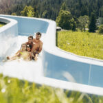 Eine vierköpfige Familie fährt an einem sonnigen Tag gemeinsam auf einer blauen Wasserrutsche, die von Gras und Bäumen umgeben ist.