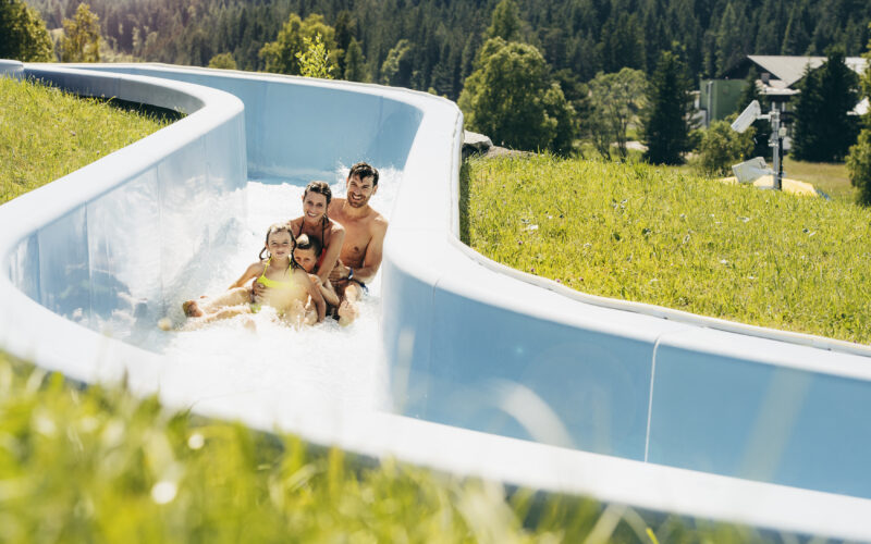Eine vierköpfige Familie fährt an einem sonnigen Tag gemeinsam auf einer blauen Wasserrutsche, die von Gras und Bäumen umgeben ist.