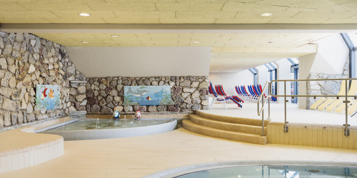 Innenpoolbereich mit runden Becken, Steinwänden, verspielter Fischdekoration und gestreiften Liegestühlen an großen Fenstern. Stufen führen von den Pools zum Sitzbereich.