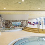 Innenpoolbereich mit runden Becken, Steinwänden, verspielter Fischdekoration und gestreiften Liegestühlen an großen Fenstern. Stufen führen von den Pools zum Sitzbereich.