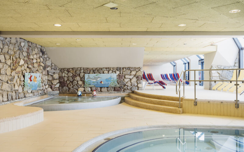 Innenpoolbereich mit runden Becken, Steinwänden, verspielter Fischdekoration und gestreiften Liegestühlen an großen Fenstern. Stufen führen von den Pools zum Sitzbereich.