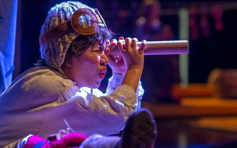 Eine Person, die eine Strickmütze und eine Brille trägt, blickt durch eine Pappröhre, als ob sie ein Teleskop benutzen würde, wobei das Bühnenlicht ihr Gesicht hervorhebt.