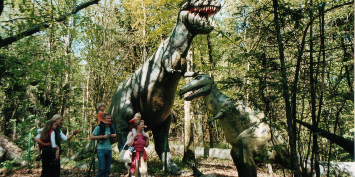 Eine Gruppe von Menschen, darunter auch Kinder, steht in der Nähe von zwei großen Dinosaurier-Statuen in einem bewaldeten Außenbereich.