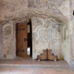 Raum mit mittelalterlicher Wandmalerei, Holztür, Ziegelfußboden und religiösen Statuen auf beiden Seiten des Eingangs.