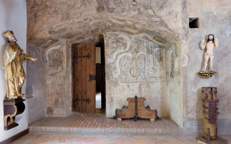 Raum mit mittelalterlicher Wandmalerei, Holztür, Ziegelfußboden und religiösen Statuen auf beiden Seiten des Eingangs.