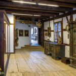 Ein Flur in einem historischen Museum ist mit Holzböden, antiken Möbeln, Glasvitrinen und verschiedenen Kunstwerken an den Wänden ausgestattet.
