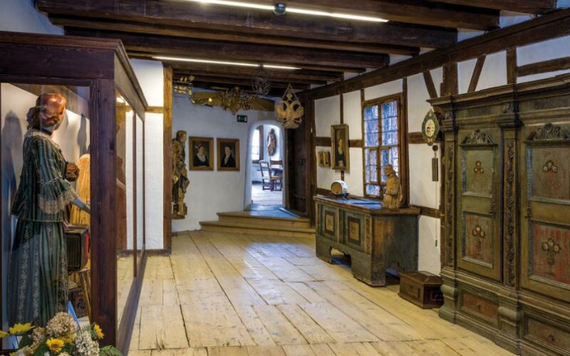 Ein Flur in einem historischen Museum ist mit Holzböden, antiken Möbeln, Glasvitrinen und verschiedenen Kunstwerken an den Wänden ausgestattet.