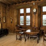 Holzgetäfelter Raum mit Bleiglasfenstern, geschnitztem Holztisch und Stühlen, einer Uhr an der Wand, religiösen Statuen und einer Truhe auf einem Holzboden.