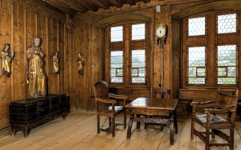 Holzgetäfelter Raum mit Bleiglasfenstern, geschnitztem Holztisch und Stühlen, einer Uhr an der Wand, religiösen Statuen und einer Truhe auf einem Holzboden.