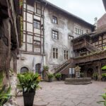 Ein historischer Steinhof mit Topfpflanzen, einem zentralen Brunnen, Holzbalkonen und Treppen, umgeben von Fachwerkwänden und Fenstern.